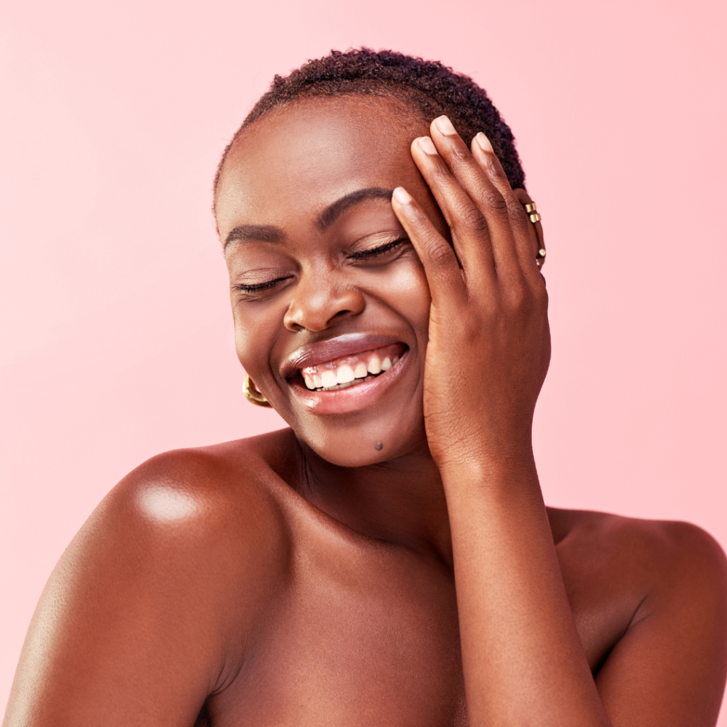 Black woman with left hand to her face lovingly with radiant skin on a pink background representing spring skin care blog by SOLEILRA 